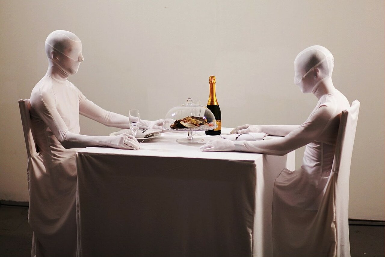 Two people in white fuull body suits sitting at a set dining table.