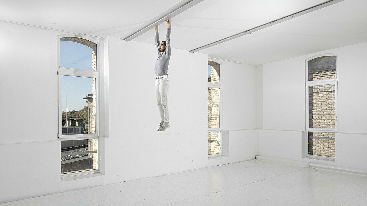 Imre Vass hanging on a beam on the ceiling of a white room.